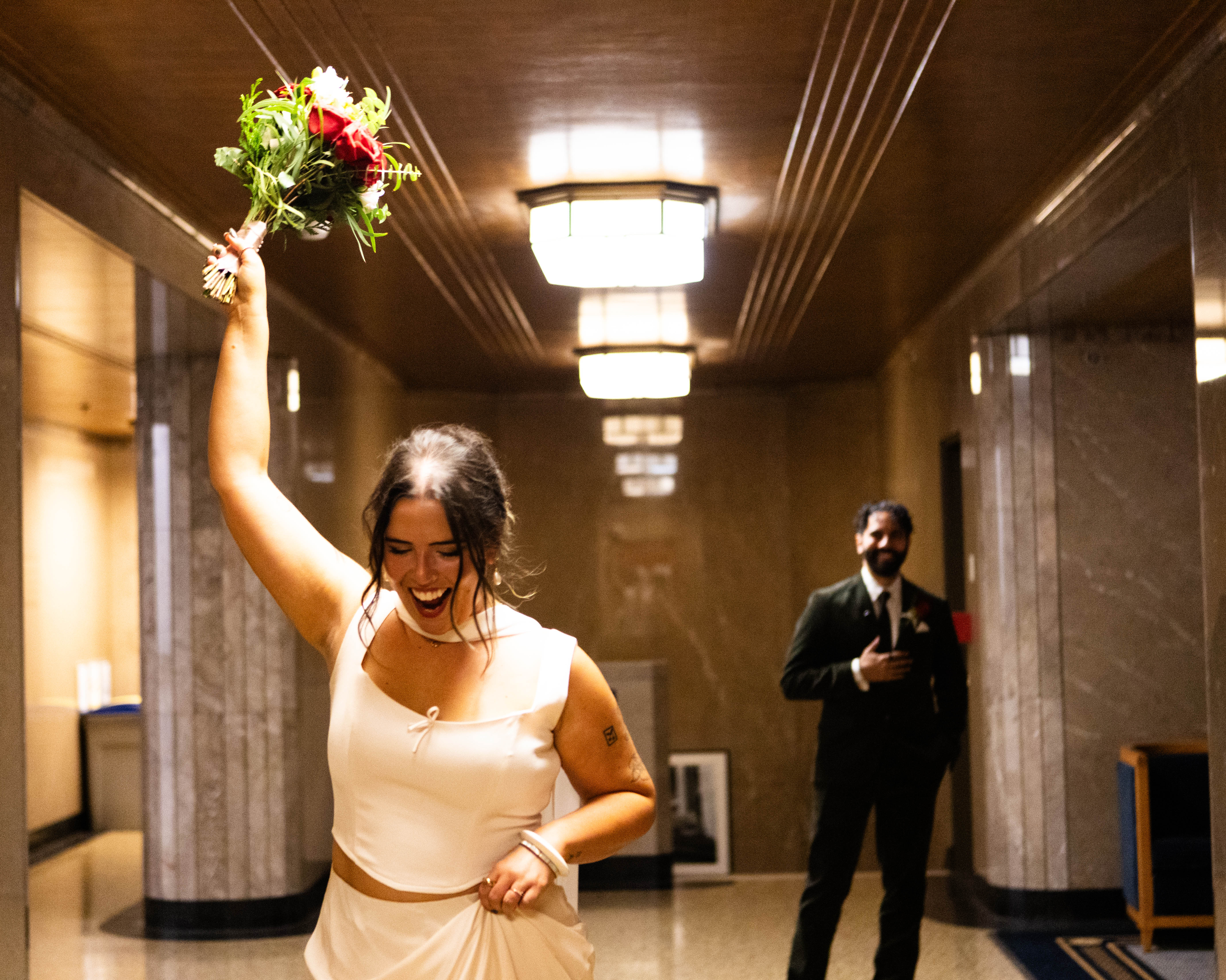 Bride raising her bouquet while walking through a warmly lit interior with the groom behind her