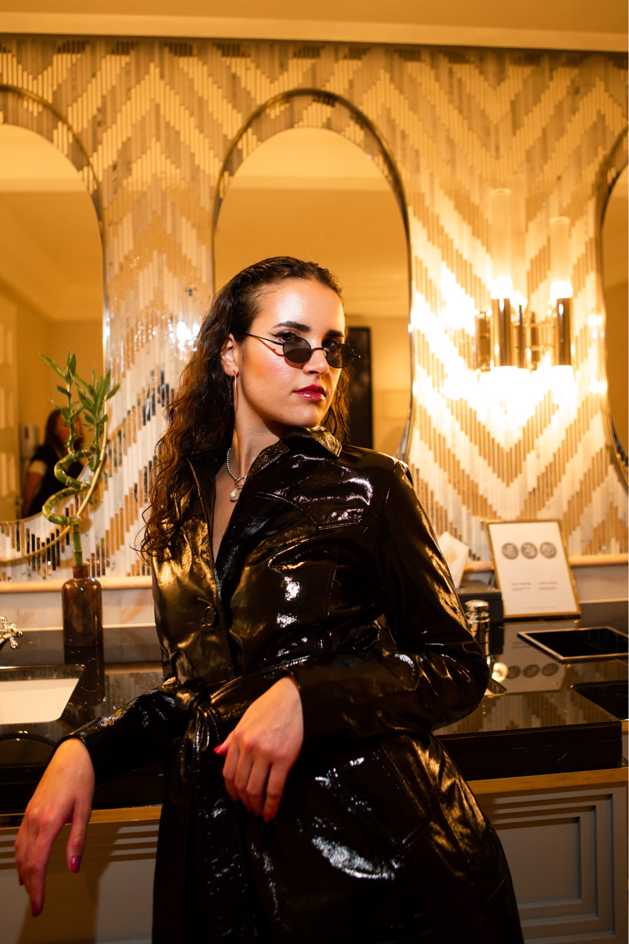 Editorial portrait of a woman in a glossy black outfit posed in a warm mirrored interior