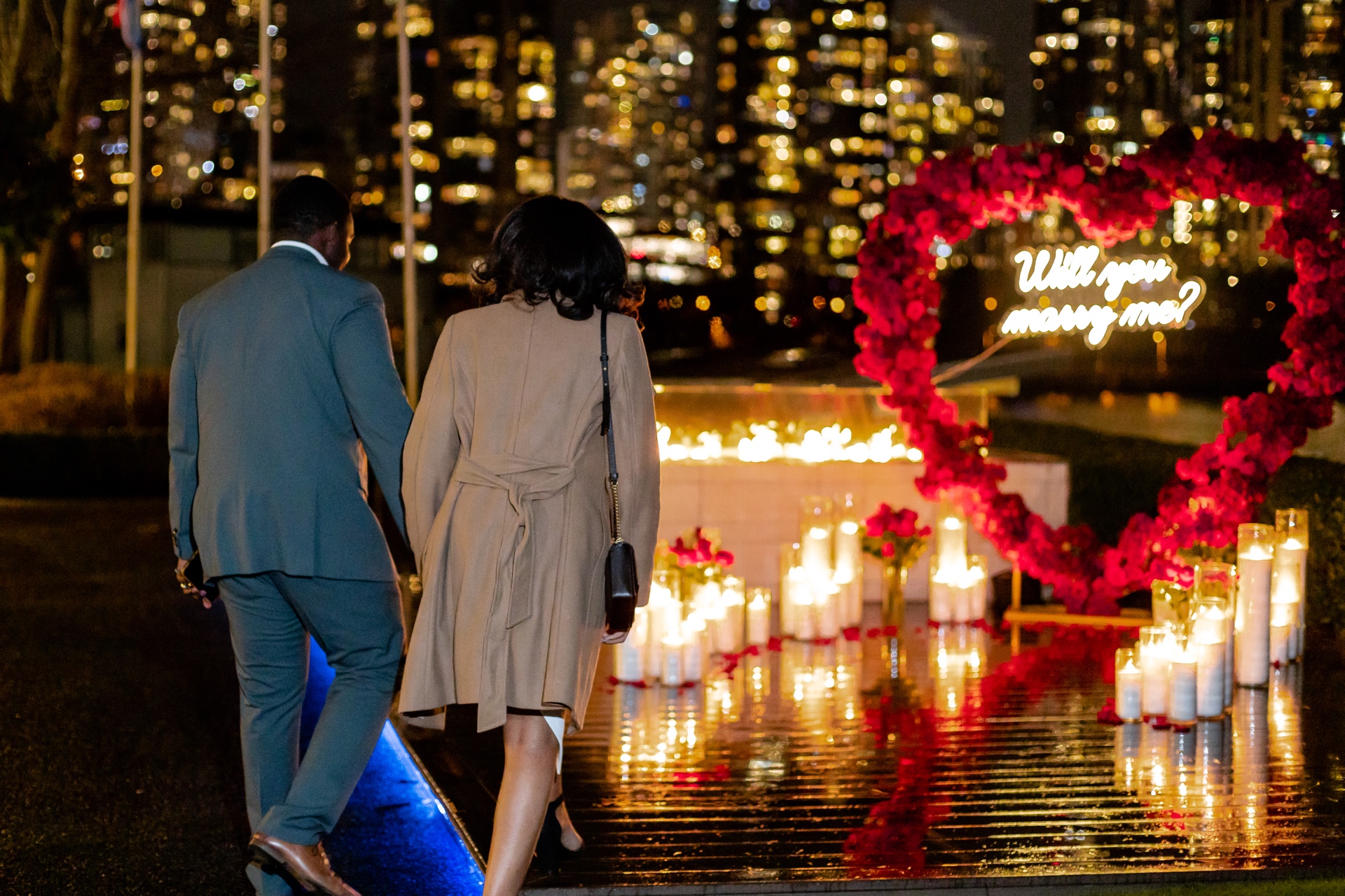 Couple walking toward a candlelit heart-shaped floral setup against a city skyline at night