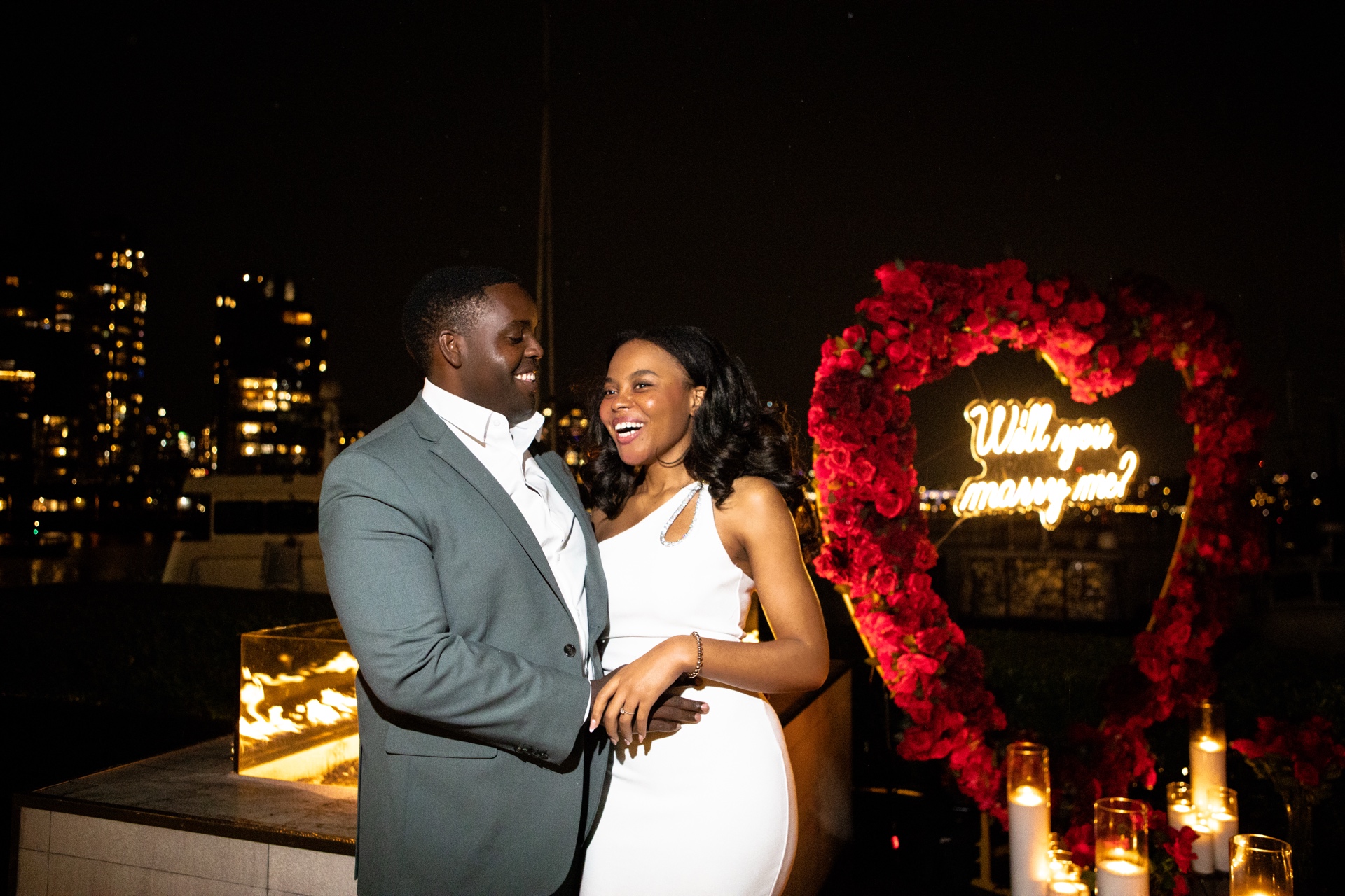 Smiling couple standing beside a glowing floral heart installation on a rooftop at night