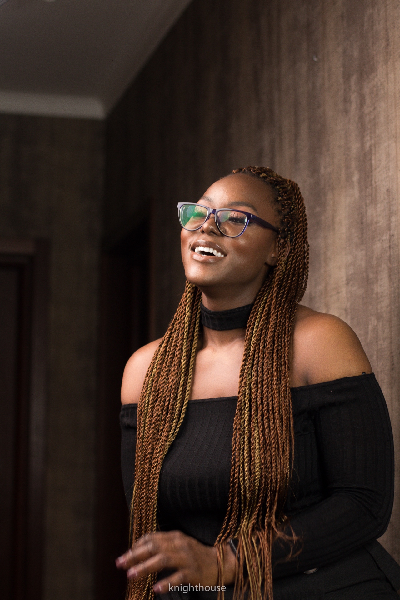 Portrait of a smiling woman with long braids and glasses wearing a black dress indoors